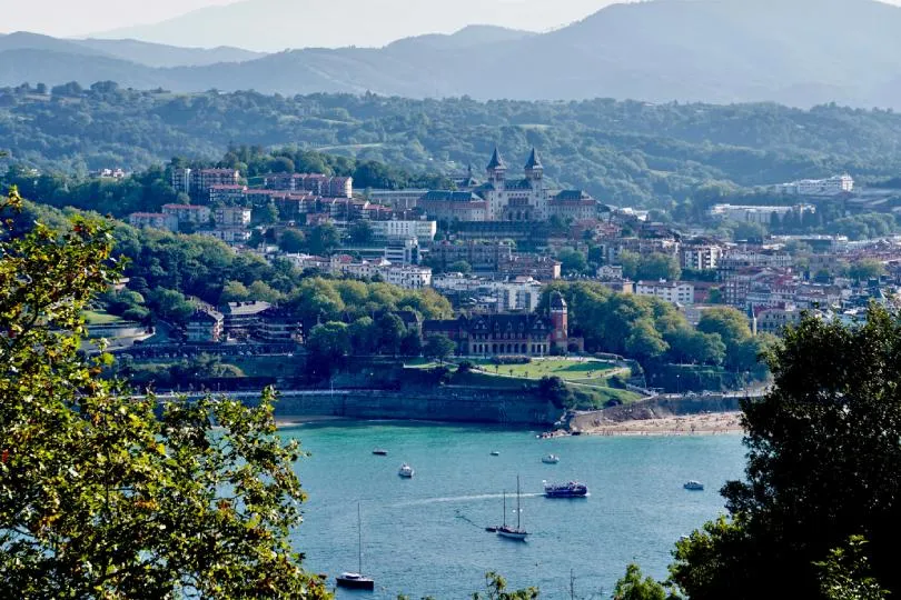 Küstenstadt mit Bucht, Segelbooten im Wasser und dicht bebauten Hügeln; im Hintergrund ein großes Gebäude mit zwei Türmen, dahinter bewaldete Berge.