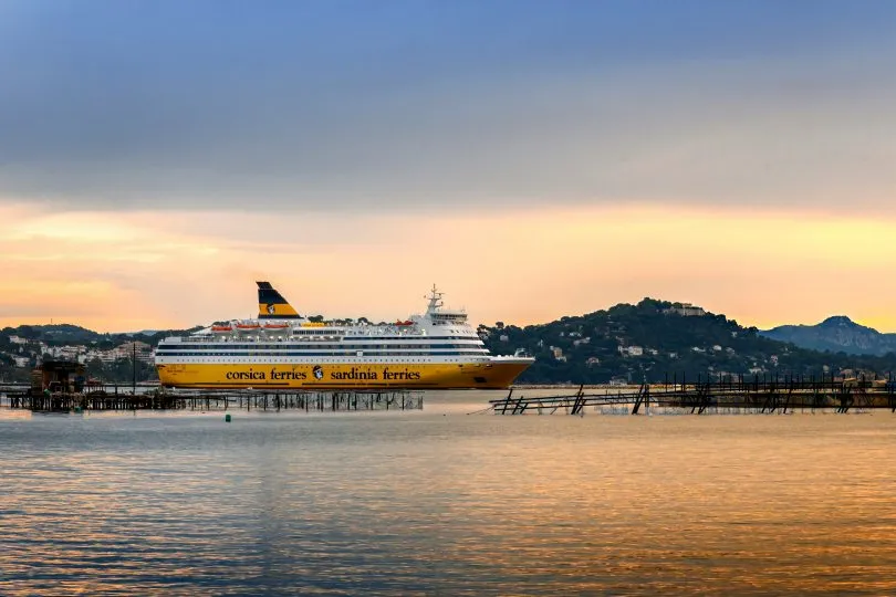 Large Corsica–Sardinia passenger ferry sailing near a harbor at sunset with wooden piers in the foreground and hills in the background.