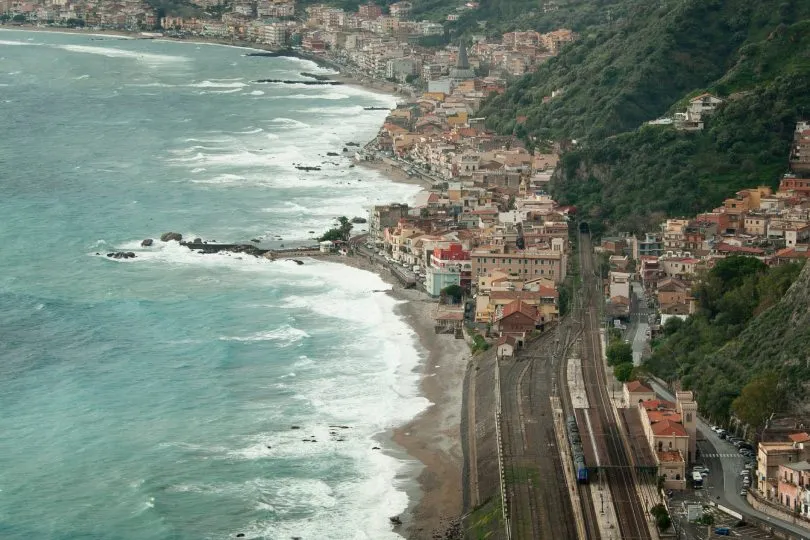 Küstenstadt in Kalabrien mit Bahnstrecke direkt am Meer: Wellen schlagen an den Strand, während Gleise und ein Bahnhof entlang der Küste durch die dicht bebaute Stadt verlaufen.