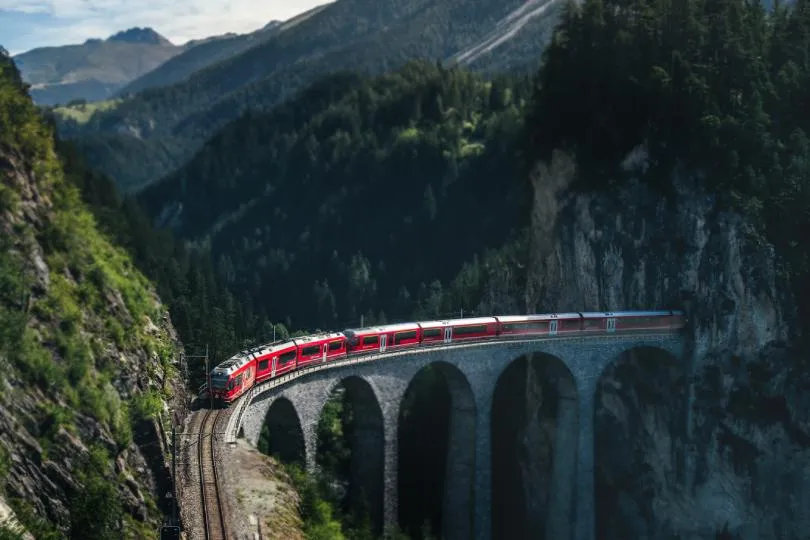 Roter Personenzug fährt über eine gebogene Steinviadukt-Brücke über eine Schlucht in einer bewaldeten Berglandschaft.