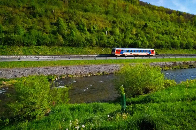 Regionalzug fährt entlang einer Bahnstrecke neben einem Fluss durch eine grüne Landschaft mit Wiesen und dicht bewaldeten Hügeln im Hintergrund.