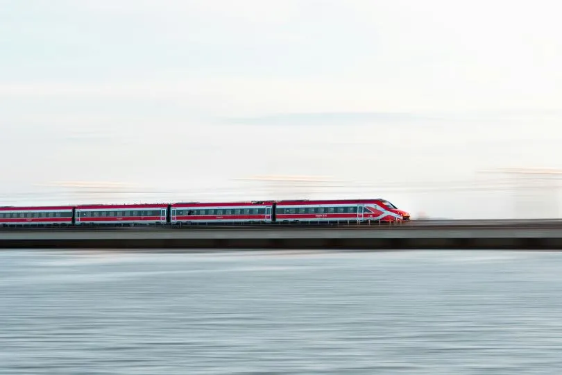 Ein rot-weißer Hochgeschwindigkeitszug fährt über eine lange Brücke oder einen Damm über dem Wasser. Durch eine lange Belichtungszeit wirkt der Zug leicht verschwommen, während Himmel und Wasser in weichen, ruhigen Farben erscheinen.