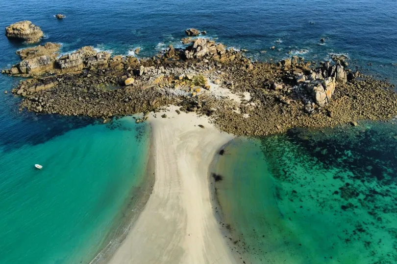Luftaufnahme einer schmalen Sandbank, die zu einer kleinen felsigen Insel im Meer führt. Das Wasser auf beiden Seiten schimmert in türkisfarbenen und dunkleren Blautönen.