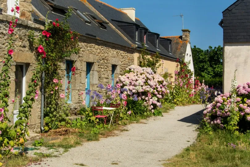 Schmale Dorfstraße mit alten Steinhäusern, geschmückt mit vielen bunten Blumen und blühenden Sträuchern entlang der Fassaden.