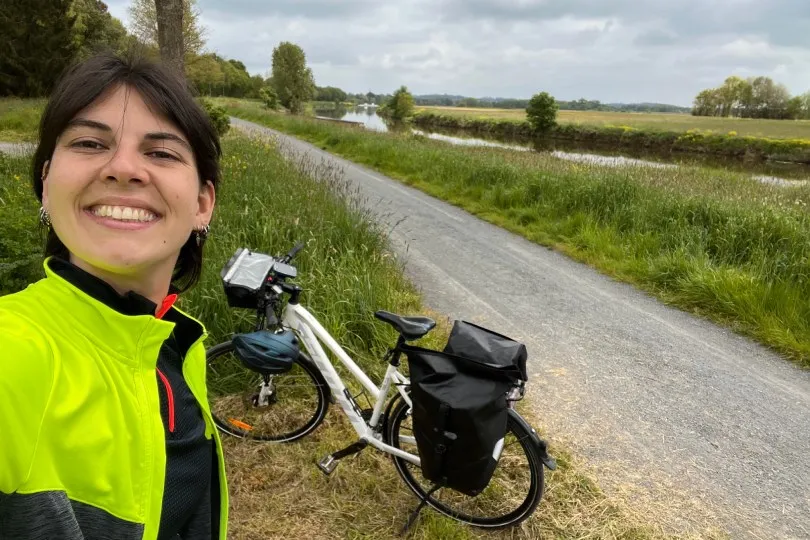 Unsere Fahrradreise-Expertin Johanne macht ein Selfie auf einem ruhigen Radweg in der Bretagne. Neben ihr steht ein bepacktes Fahrrad, während sich im Hintergrund eine idyllische Landschaft mit Wiesen, einem Fluss und bewölktem Himmel erstreckt.