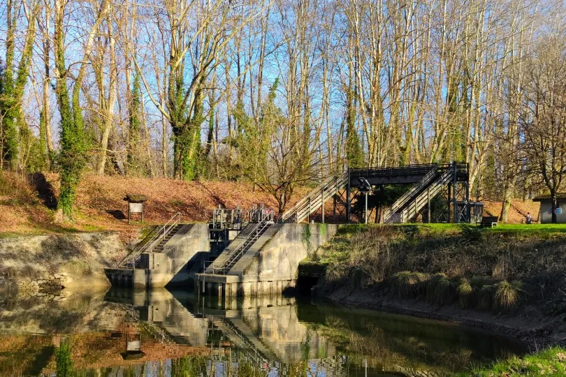 Eine kleine Schleuse an einem ruhigen Kanal, umgeben von Bäumen, mit einer Holzbrücke und klarer Spiegelung im Wasser.