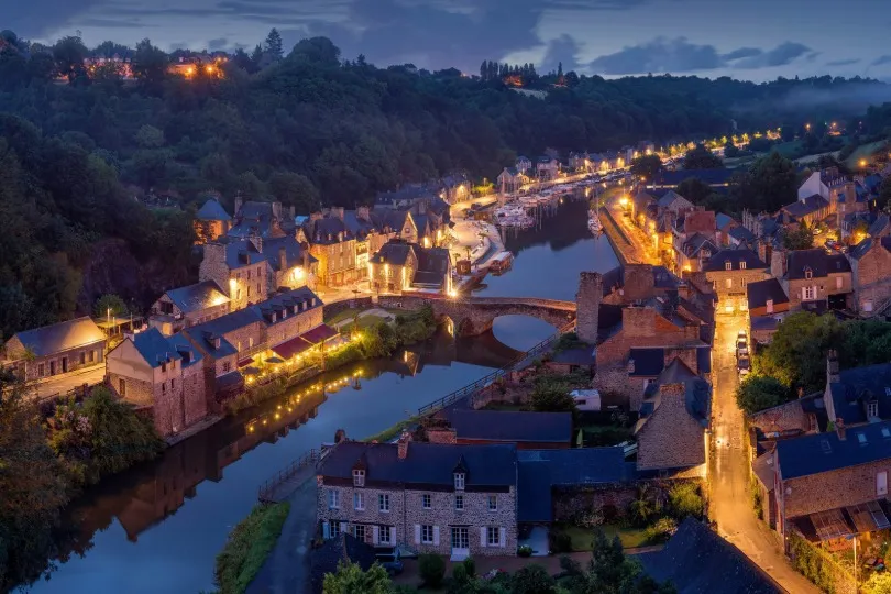 Eine stimmungsvolle Abendaufnahme der Altstadt von Dinan in der Bretagne mit beleuchteten Fachwerkhäusern, einer Steinbrücke über den Fluss und warmem Licht entlang der Uferpromenade.