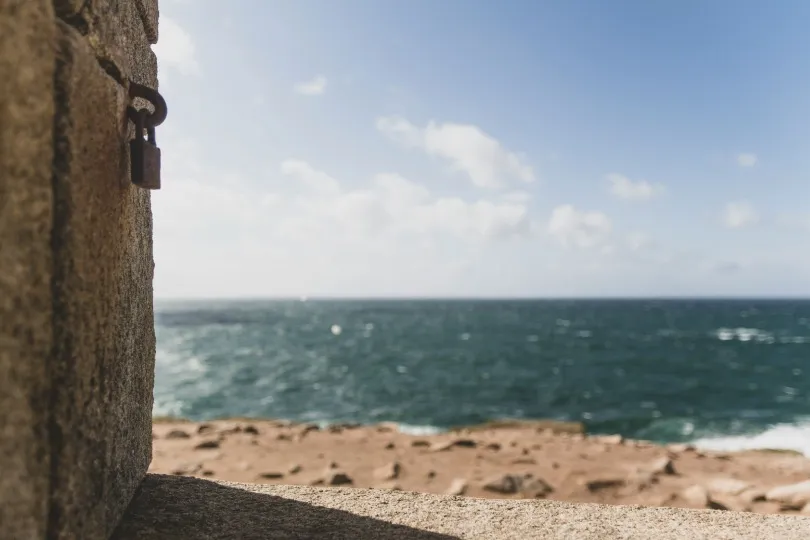 Blick durch eine Steinöffnung auf das Meer, mit Sandstrand, Wellen und blauem Himmel im Hintergrund.