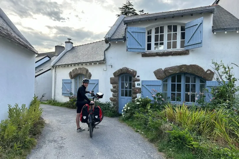 Eine Person mit Fahrrad hält auf einer schmalen Straße vor einem charmanten, weiß gestrichenen Haus mit blauen Fensterläden und blühendem Garten in einem ruhigen Dorf.