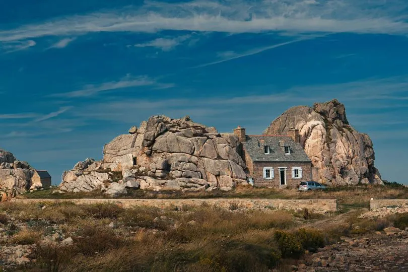 Steinhaus an der bretonischen Granitküste in Plougrescant, eingebettet zwischen großen rosa Granitfelsen unter blauem Himmel.