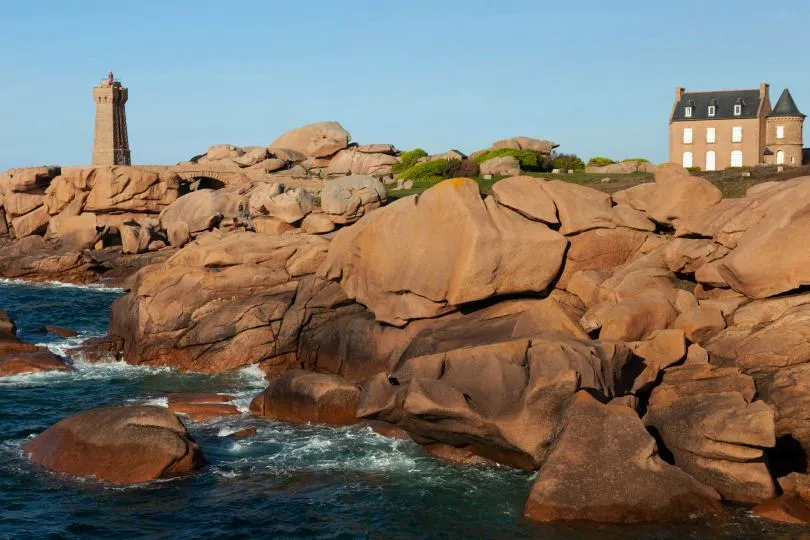 Rosa gefärbte Granitfelsen an einer zerklüfteten Küste ragen aus dem Meer, umspült von Wellen. Auf einem Felsplateau steht ein Leuchtturm neben einer kleinen Steinbrücke, weiter rechts ein einzelnes Haus mit Turm. Die Szene ist von warmem Licht beleuchtet, unter klarem Himmel.