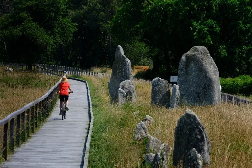 Eine Person fährt mit dem Fahrrad auf einem Holzweg entlang großer, aufrecht stehender Steine in einer grünen Landschaft.