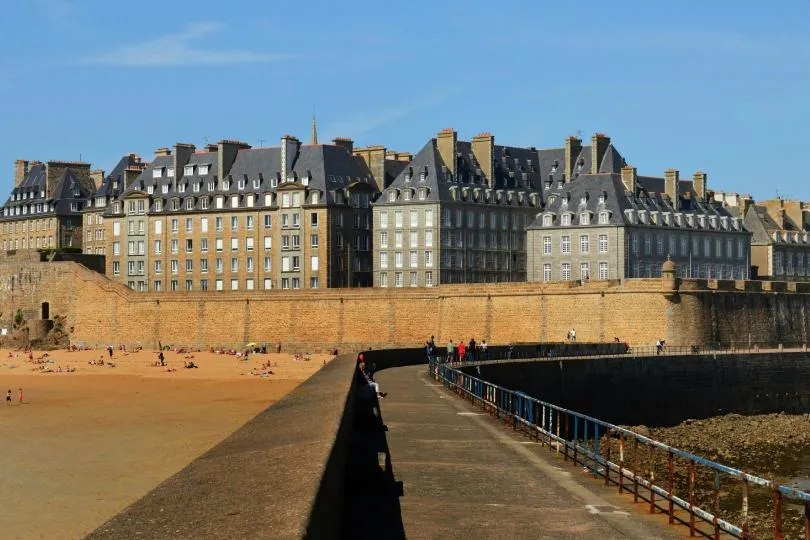 Historische Küstenstadt mit hohen steinernen Festungsmauern direkt am Meer. Dahinter stehen dicht gedrängt mehrstöckige Gebäude mit grauen Schieferdächern und vielen Schornsteinen. Im Vordergrund führt ein schmaler Pier mit Geländer entlang der Mauer.