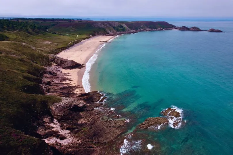 Alt-Text (Deutsch):  Luftaufnahme einer geschwungenen Bucht mit türkisfarbenem Wasser und hellem Sandstrand. Felsige Klippen mit grün bewachsenen Hügeln rahmen die Küste ein. Ein schmaler Wanderweg verläuft entlang der Steilküste, und vereinzelt sind kleine Menschen am Strand zu erkennen. Das Meer ist ruhig, und der Himmel leicht bewölkt.