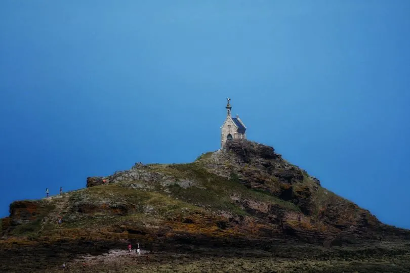 Kleine Kapelle mit Kreuz auf der Spitze steht auf einem felsigen Hügel direkt am Meer. Der Hügel ist von rauem, unebenem Gestein geprägt, mit wenigen Grasflächen. Einige kleine Menschen sind am Hang zu erkennen und wirken im Vergleich zur Kapelle sehr klein. Der Himmel ist klar und intensiv blau, ohne Wolken.