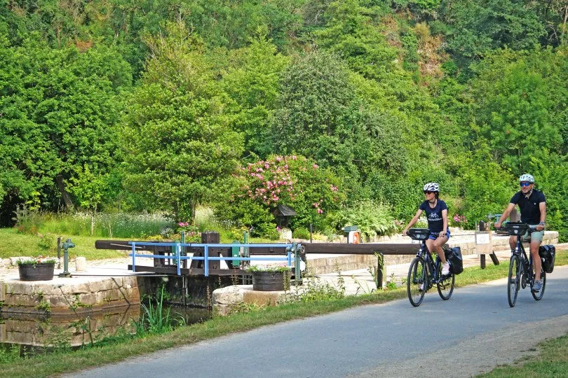 Zwei Radfahrer mit Helmen fahren auf einem ruhigen Weg neben einem kleinen Kanal mit Schleuse, umgeben von üppigem Grün, Bäumen und blühenden Pflanzen an einem sonnigen Tag.