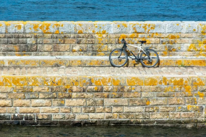 Ein Fahrrad lehnt an einer steinernen Mauer mit gelben Flechten direkt am Meer.