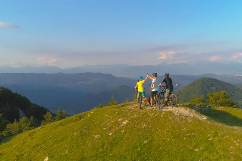 Drei Radfahrer stehen auf einem Hügel und blicken auf eine weite Berglandschaft.