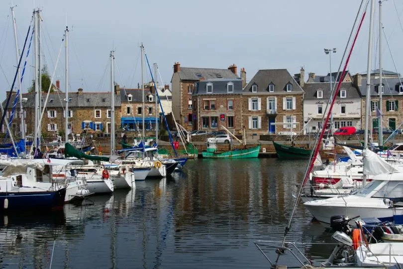 Kleiner Hafen mit zahlreichen Segelbooten, deren Masten dicht nebeneinander in den Himmel ragen. Im ruhigen Wasser spiegeln sich die Boote und Gebäude. Im Hintergrund stehen typische steinerne Häuser mit grauen Dächern, Cafés und Geschäften entlang der Uferpromenade. Die Szene wirkt wie ein ruhiges Fischerdorf an der Küste.