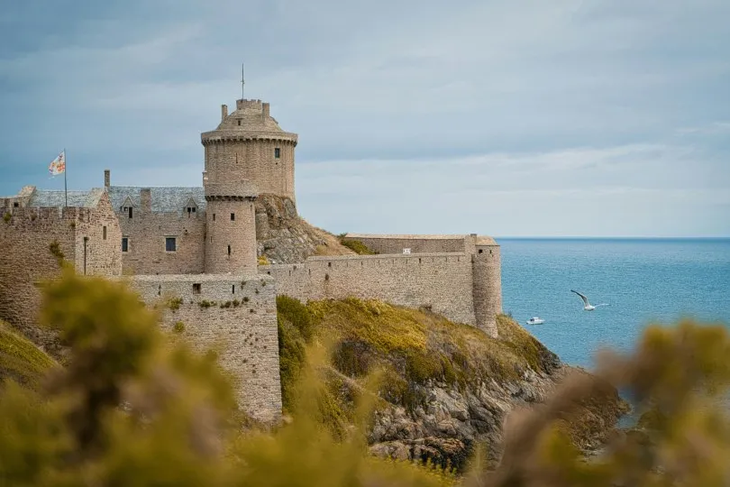 Steinerne Festung (Fort La Latte) auf einer Klippe am Meer, mit Türmen und Mauern über der Küste, unter bewölktem Himmel.