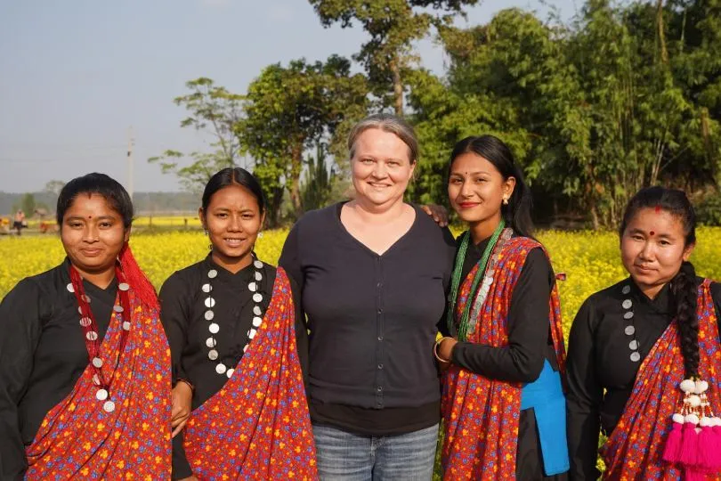 Fünf Frauen stehen gemeinsam vor einem gelben Blütenfeld; vier tragen traditionelle Kleidung, eine ein dunkles Oberteil und Jeans.