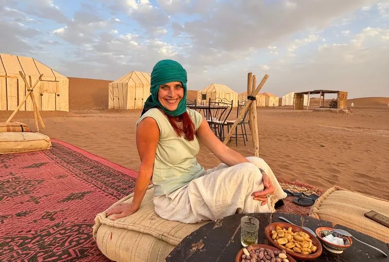 Touristin mit grünem Turban sitzt an einem kleinen Essenstisch und lächelt in die Kamera. Hinter ihr stehen Wüstenzelte. 