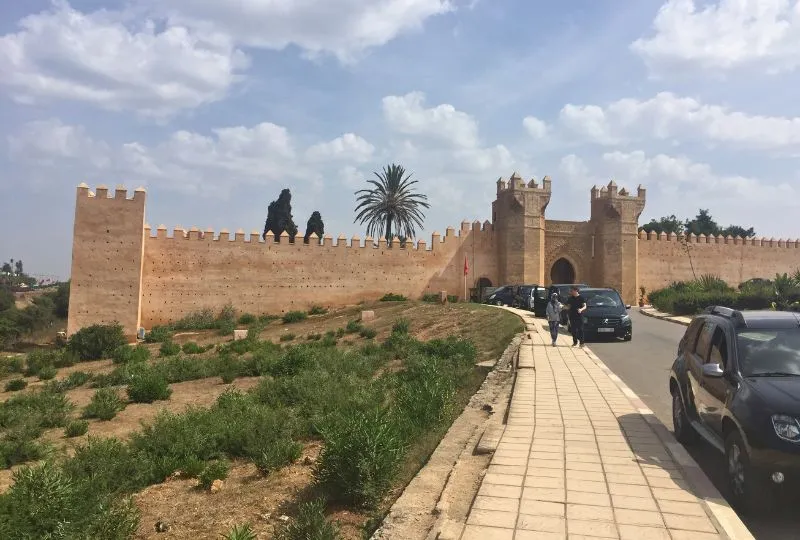 Autos stehen auf einer Straße direkt vor einer Mauer mit imposanten Türmen in Marokko.