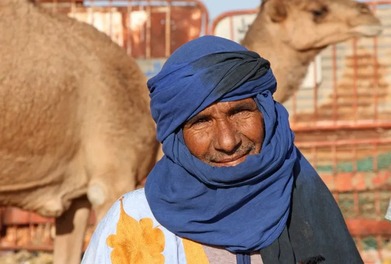 Einheimischer in Marokko mit blauem turban lächelt in die Kamera. Hinter ihm steht ein Kamel.
