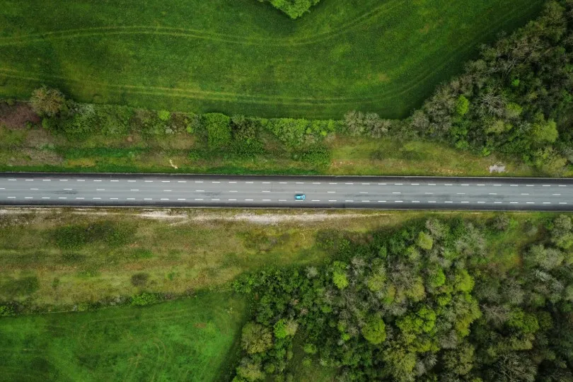 Luftaufnahme einer geraden Landstraße, auf der ein einzelnes Auto fährt, umgeben von grünen Feldern und Waldflächen.