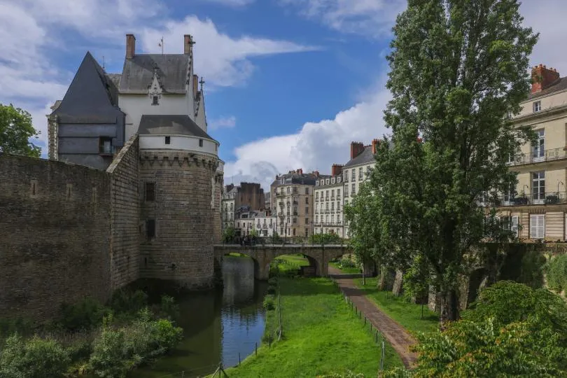 Ein historisches Stadttor mit runder Wehranlage aus Stein liegt am Ufer eines ruhigen Flusses. Eine kleine Brücke führt in die Altstadt mit klassischen französischen Gebäuden, während ein grüner Uferweg und hohe Bäume die Szene umgeben.