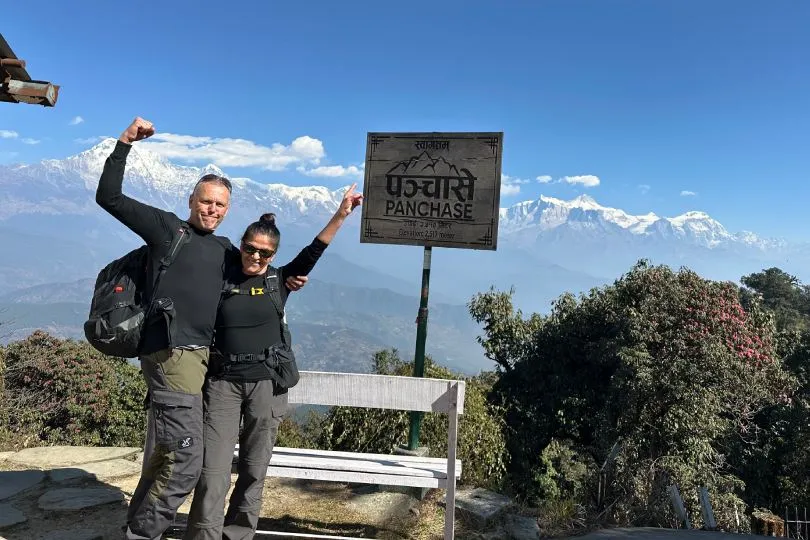 Zwei Personen stehen vor einem Schild mit der Aufschrift „Panchase“ und posieren jubelnd mit erhobenen Armen. Sie tragen Wanderkleidung und Rucksäcke. Im Hintergrund sieht man eine beeindruckende Berglandschaft mit schneebedeckten Gipfeln und blauem Himmel – vermutlich im Himalaya-Gebiet.