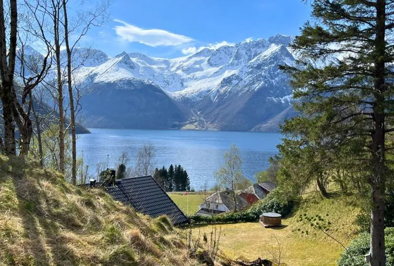Landschaft in Norwegen mit Wiesen und kleinen Häuschen im Vordergrund und einem Fjord und schneebedeckten Bergen im Hintergrund.