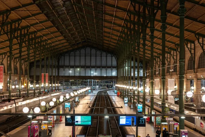 Große Bahnhofshalle mit Gleisen und Zügen am Bahnsteig, beleuchtet von vielen Lampen unter einer hohen Stahl- und Glasdachkonstruktion.