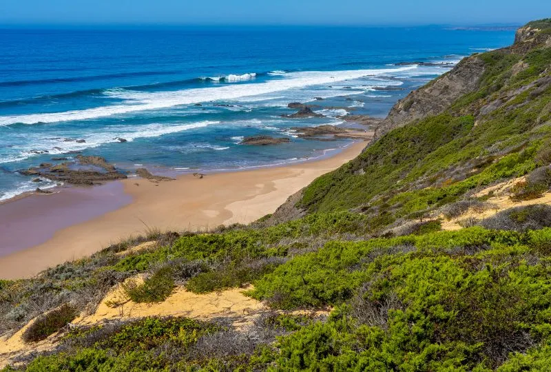 Das grüne Land trifft auf das blaue Meer am Fischerweg in Portugal. Dazwischen liegt der Sandstrand. 