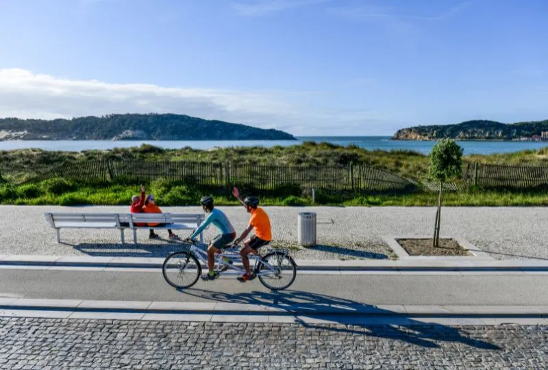 Fahrradfahrer fahren die grüne Küste in Portugal entlang. Die Locals auf einer Bank winken ihnen zu. 