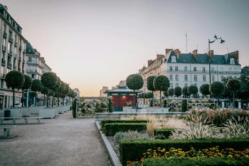 Ein gepflegter Stadtplatz in Rennes mit symmetrisch geschnittenen Bäumen, Blumenbeeten und klassischer französischer Architektur im warmen Abendlicht.
