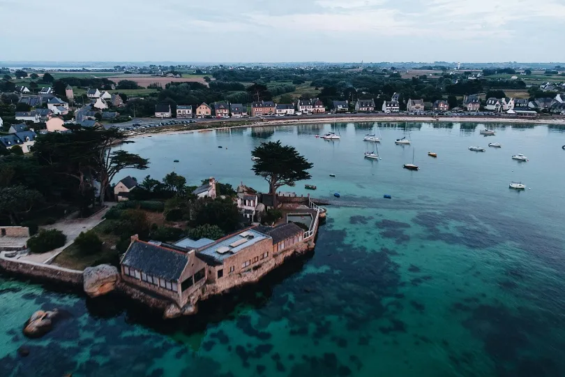 Luftaufnahme einer geschützten Bucht mit ruhigem, klarem Wasser, in dem mehrere kleine Boote vor Anker liegen. Im Vordergrund befindet sich eine kleine Landzunge mit Häusern und Bäumen, umgeben vom Meer. Entlang der gegenüberliegenden Uferlinie stehen dicht gereihte Häuser eines Küstenortes, dahinter erstrecken sich Felder und Landschaft im Landesinneren.