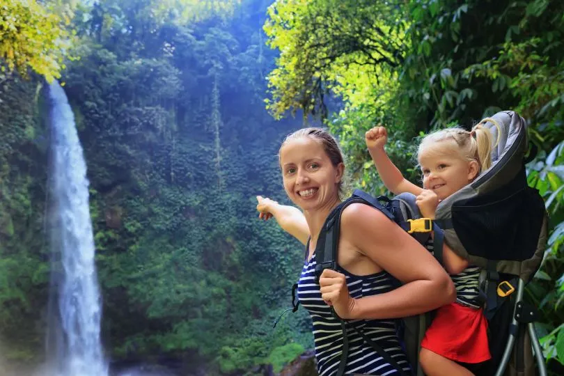 Eine Frau trägt ein Kind in einer Kraxe und zeigt auf einen Wasserfall im dichten Wald.