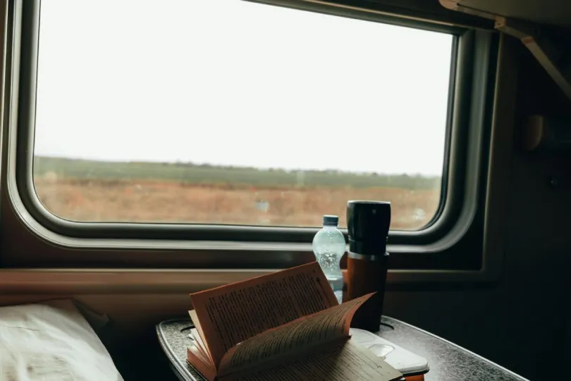 Offenes Buch und Thermosbecher auf einem Tisch im Zugabteil, mit Blick aus dem Fenster auf eine vorbeiziehende Landschaft.
