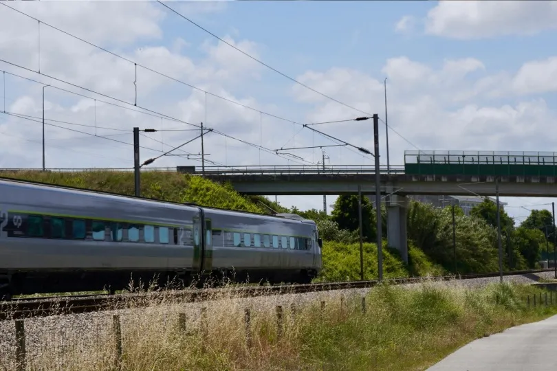Französischer Personenzug fährt auf einer Bahnstrecke durch eine grüne Landschaft, mit Brücke und Oberleitungen im Hintergrund.