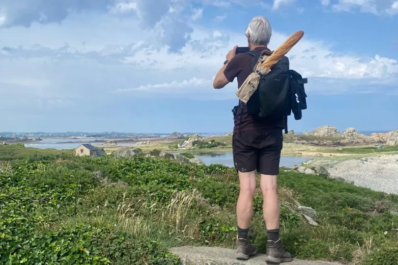 Ein Wanderer fotografiert die Küstenlandschaft der Bretagne mit Meer, Felsen und weitem Himmel.