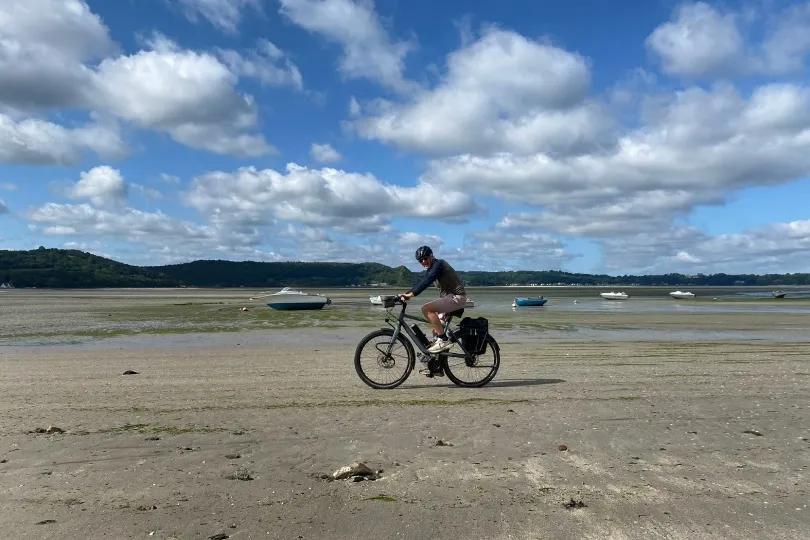 Eine Person fährt mit dem Fahrrad über einen weiten Sandstrand bei Ebbe, während im Hintergrund kleine Fischerboote im flachen Wasser liegen und sich eine hügelige Küstenlandschaft unter blauem Himmel mit Wolken erstreckt.