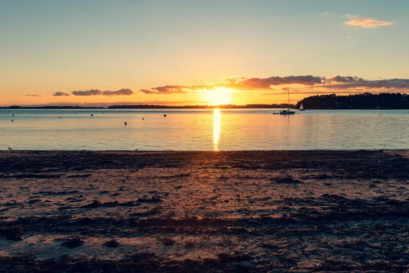 Sonnenuntergang über ruhigem Wasser, im Vordergrund ein dunkler Strand, auf dem Wasser wenige Boote und Vögel, am Horizont eine flache Küstenlinie unter leicht bewölktem Himmel.
