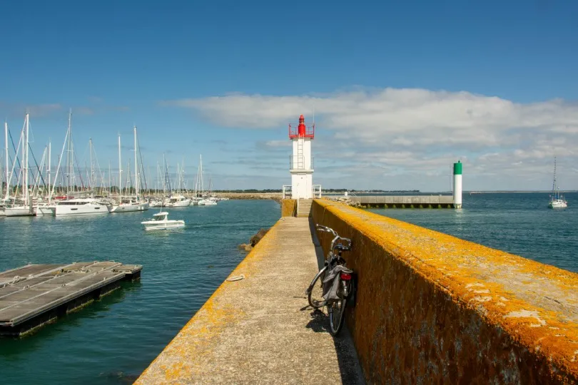 Langer Steg führt zu einem weißen Leuchtturm mit roter Spitze, daneben ein Fahrrad, links ein Hafen mit vielen Segelbooten und ruhigem Wasser unter blauem Himmel.