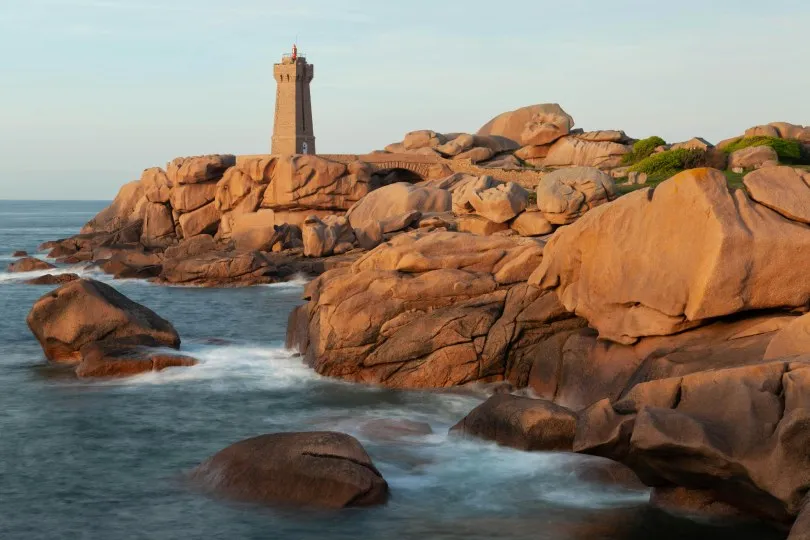 Ein Leuchtturm steht auf einer zerklüfteten rosafarbenen Granitküste am Meer. Die warm beleuchteten Felsen und das sanft bewegte Wasser erzeugen eine ruhige Küstenlandschaft.