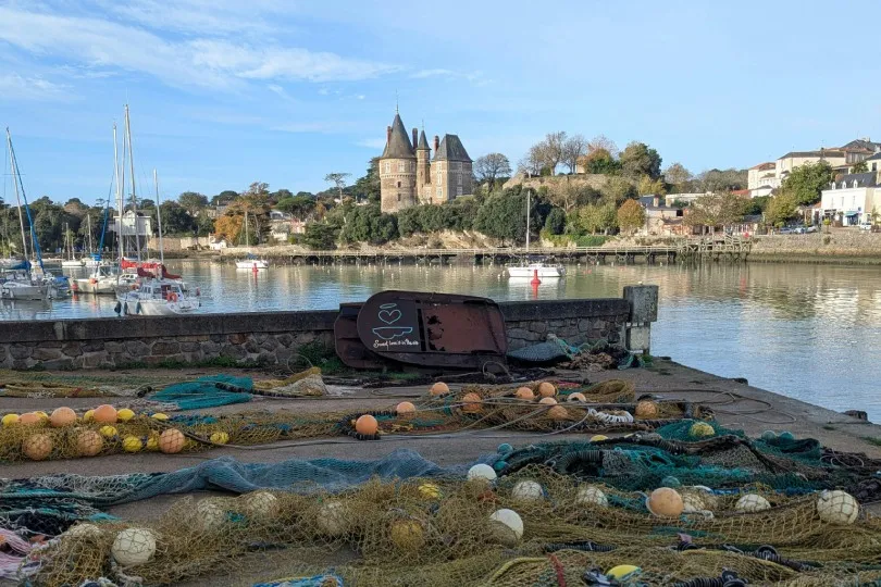 Hafen mit ruhigem Wasser, im Vordergrund Fischernetze und Bojen, dahinter ein Flussufer mit Booten und ein Gebäude mit Türmen auf einem Hügel unter blauem Himmel.