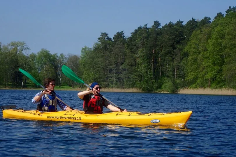 Zwei Reisende fahren mit einem Kayak über einen See, umringt von Nadelbäumen, im Baltikum.