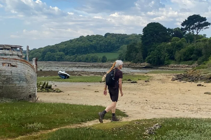 Wanderer an einer grünen Küstenlandschaft in der Bretagne mit altem Holzboot, Strand und bewaldeten Hügeln.