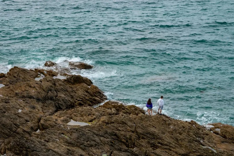 Zwei Personen stehen auf felsiger Küste in der Bretagne und blicken auf das Meer.