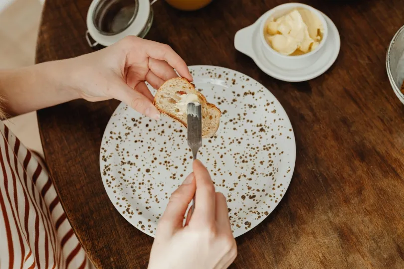 Eine Person bestreicht eine Scheibe Baguette mit Butter. Das Brot liegt über einem großen, weiß gesprenkelten Teller, während im Hintergrund eine kleine Schale mit Butter auf einem Holztisch steht.